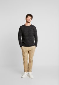Black textured sweater with ribbed design, paired with light beige slim-fit pants and white sneakers. Captured standing against a light background.