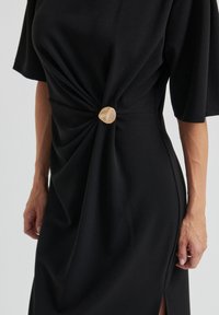 Robe noire à manches courtes avec un détail torsadé et un bouton crème à la taille ; tissu lisse avec une coupe décontractée.