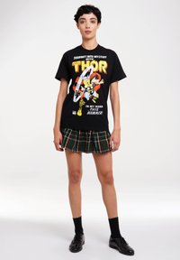 Schwarzes T-Shirt mit einem bunten Thor-Motiv; kombiniert mit einem karierten, plissierten Rock in grünen, roten und gelben Tönen; getragen mit schwarzen Schuhen und Socken.