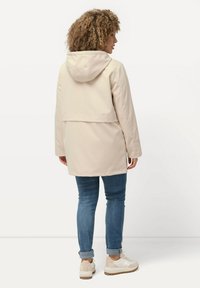 Giacca impermeabile color beige chiaro con cappuccio, design di lunghezza media e dettaglio di ventrazione sul retro, abbinata a jeans blu arrotolati e sneakers bianche.