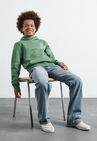 Felpa verde in cotone con tasca frontale e logo, abbinata a jeans azzurri chiari e scarpe bianche senza lacci, seduta su una sedia.