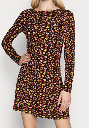Kobieta w czarnej mini sukience z długim rękawem ozdobionej kolorowym, drobnym kwiatowym wzorem, z ręką na biodrze, na jasnym, jednolitym tle.