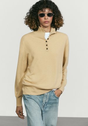 Vrouw met krullend haar die kleine ovale zonnebrillen draagt, een beige vest met knopen en lichtblauwe spijkerbroek met één hand in de zak.