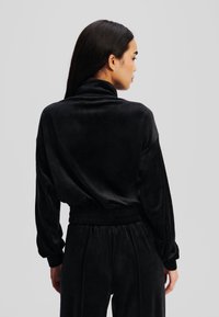 Melnas samta treniņtērps ar augstu apkakli, brīvu piegriezumu un savilktu jostu. Garas piedurknes ar aprocēm un smalka tekstūra visā audumā.