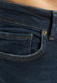 Nærbilde av mørkeblå denimjeanslomme med synlige sømmer, metallnagle og del av linningen mot bar hud.