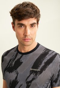 Camiseta gris con patrón de pinceladas negras, cu neckline negro redondo y pequeño logo en el pecho. Material de algodón suave, mangas cortas.