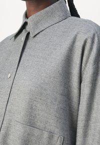 Chemise grise à manches longues, en tissu texturé, avec col classique et une poche poitrine. Caractéristiques : coupe décontractée et coutures soignées.