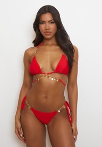 Conjunto de bikini rojo que presenta copas triangulares y lazos laterales, acentuado con un hardware circular dorado y un detalle de cadena alrededor de la cintura.