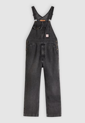 Salopette en denim noir avec bretelles réglables, poches avant, fermetures latérales à boutons et une étiquette en patch sur la poche poitrine.