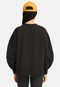 Schwarzer, übergroßer Sweatshirt mit gesenkten Schultern, gerippten Bündchen und Saum. Kombiniert mit einer gelben Kordmütze, die einen verstellbaren Riemen hinten hat.