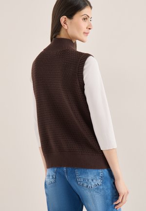 Femme portant un gilet sans manches à col roulé en tricot marron foncé sur un haut blanc et un jean bleu, vue de profil sur un fond beige.