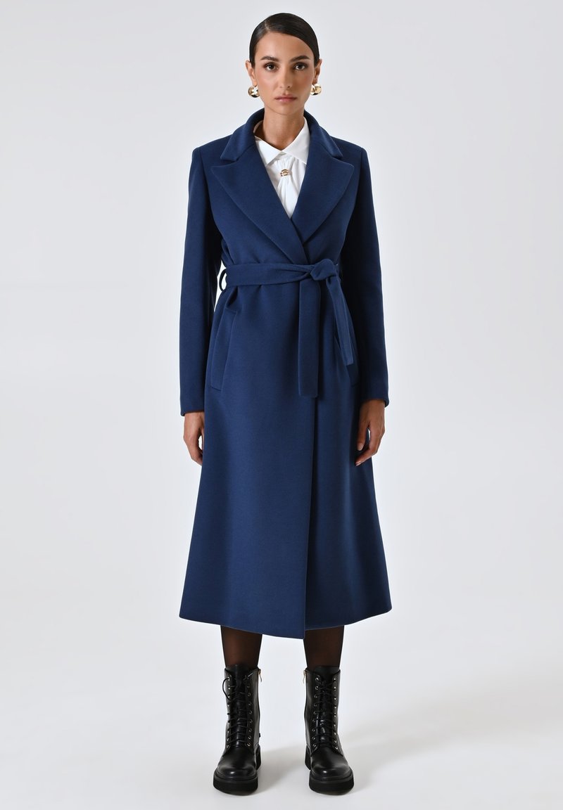 Cappotto avvolgente in misto lana blu navy con vita cintrata, colletto a punta e maniche lunghe. Abbinato a stivali neri alla caviglia con lacci.
