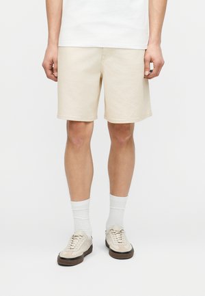 Uomo con pantaloncini beige fino al ginocchio, calzini bianchi e scarpe da ginnastica beige, in piedi davanti a uno sfondo bianco uniforme.