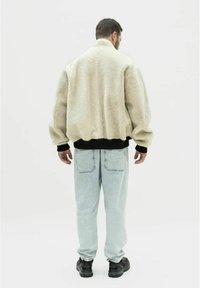 Faux fur jakke i creme, oversized design med sænkede skuldre, sort ribbet kant og manchetter, båret over lyseblå jeans og sorte sneakers.