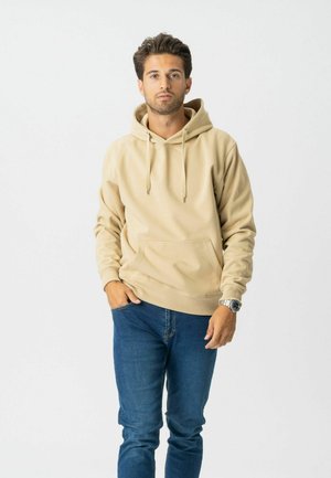 Mand med mørkt hår iført beige hættetrøje, blå jeans og armbåndsur, stående med den ene hånd i lommen mod en ensfarvet baggrund.