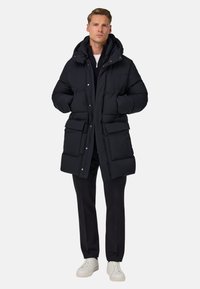 Černá péřová bunda s kapucí, se zipem, velkými předními kapsami a prošívanou strukturou. Spárována s tmavými kalhotami a bílými teniskami.