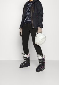 Skiausrüstung mit einer schwarzen wasserdichten Jacke, einem grafischen Pullover, schwarzen Hosen, grauen Handschuhen und schwarzen Skistiefeln, die einen weißen Helm hält.