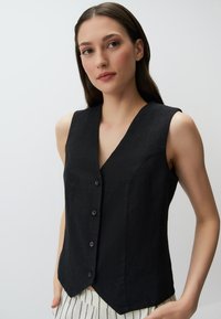 Gilet sans manches noir avec un décolleté en V profond, doté de six boutons sur le devant, en tissu texturé et avec un ourlet légèrement incurvé. Porté par-dessus un bas rayé.
