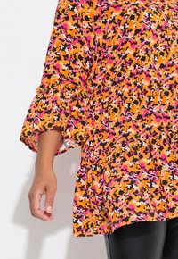 Bunte Bluse mit schichtenförmigen, rüschigen Glockenärmeln. Der Stoff hat ein abstraktes Camouflage-Muster in Orange, Pink, Schwarz und Weiß.