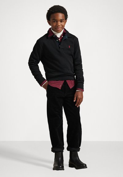 Črna sweatshirt, prepleskana čez rdečo karirano srajco, v kombinaciji s črnimi kordnimi hlačami in črnimi čevlji. Urejene, priložnostne plasti z strukturirano obliko.