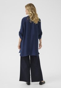 Tumši zila, viegla, krunkaina auduma krekls ar sarullētām piedurknēm, apvienots ar plašu tumšo džinsu biksēm, kam ir taisna griezuma un sānu šķēlumi.