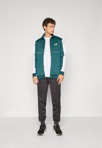Teal treningsjakke med hvite ermer, glidelås og logo. Kombinert med tettsittende grå joggebukser og svarte sneakers.