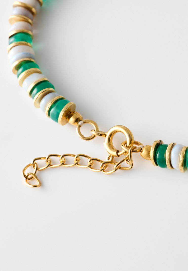 Goldfarbener Kettenanhänger mit grünen und weißen runden Perlen sowie einem Verschluss. Merkmale sind abwechselnd glänzende und matte Oberflächen sowie Details in der Hardware.