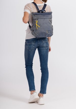 Kvinde i hvide sneakers og blå jeans iført en stor grå rygsæk med gul lynlås, stående mod en ensfarvet lys baggrund.