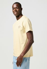 Camiseta de algodón amarilla de manga corta con cuello redondo, que presenta un logo bordado en el lado izquierdo del pecho y un corte relajado.