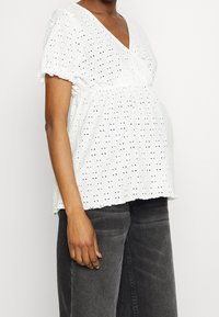 Vit, kortärmad, v-ringad topp i texturerad tyg med små utskärningar. Låtande passform, designad för graviditet, matchad med mörka jeans.