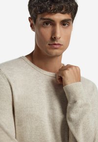 Beżowy sweter dzianinowy z okrągłym dekoltem i ściągaczami, charakteryzujący się miękką fakturą. Modelka trzyma kołnierz z neutralnym wyrazem twarzy.