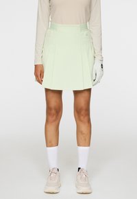 Gonna da golf verde chiaro con pieghe, realizzata in tessuto morbido. Abbinata a una maglietta beige a maniche lunghe e calze bianche, con sneakers rosa chiaro.
