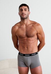 Boxer in grigio con una fascia nera etichettata 'HUGO.' Modello con capelli corti e fisico ben definito, in piedi contro uno sfondo neutro.