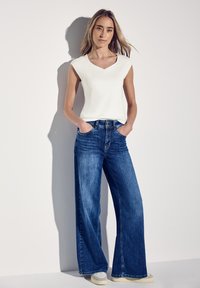 Top bianco senza maniche con scollatura a V, abbinato a jeans in denim blu scuro a gamba larga. Il modello sta in piedi con le mani nelle tasche davanti a uno sfondo semplice.