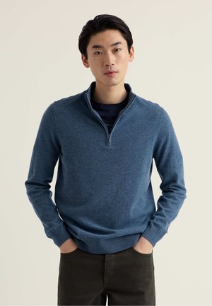 Niebieski sweter z zamkiem przy szyi, wykonany z miękkiej, teksturowanej tkaniny. Długie rękawy, ściągacze na mankietach i dole, w połączeniu z ciemnymi spodniami. Minimalistyczny design.