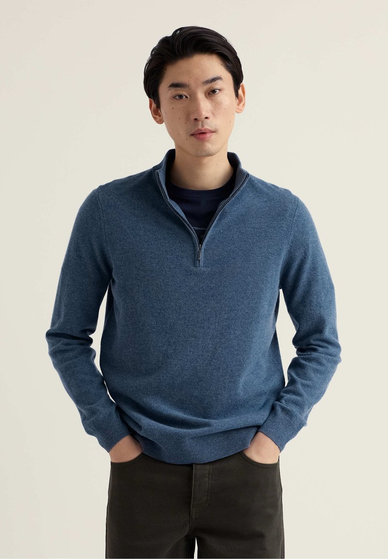 Pull bleu à col zippé en tissu doux et texturé. Manches longues, poignets et ourlet côtelés, associé à un pantalon sombre. Design minimaliste.