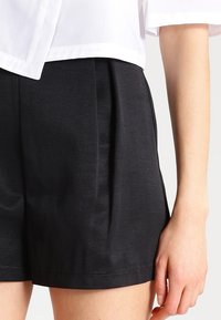 Czarne, dopasowane szorty o gładkiej fakturze, z bocznymi plisami i płaskim paskiem w pasie, zestawione z białą, krótką bluzką.