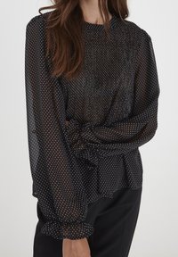 Blusa nera trasparente con pois bianchi, maniche a sbuffo e polsini arricciati. Il tessuto è leggero e fluido. Scollo semplice e rotondo.