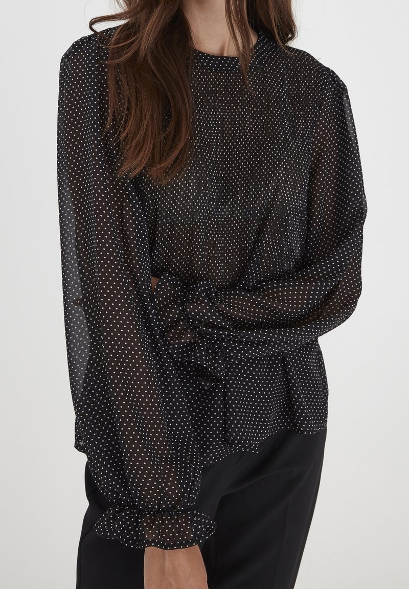 Blusa nera trasparente con pois bianchi, maniche a sbuffo e polsini arricciati. Il tessuto è leggero e fluido. Scollo semplice e rotondo.