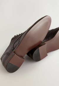 Chaussures à talons marron en cuir texturé, ornées de perforations décoratives et d'une semelle en caoutchouc plate avec des détails rayés.