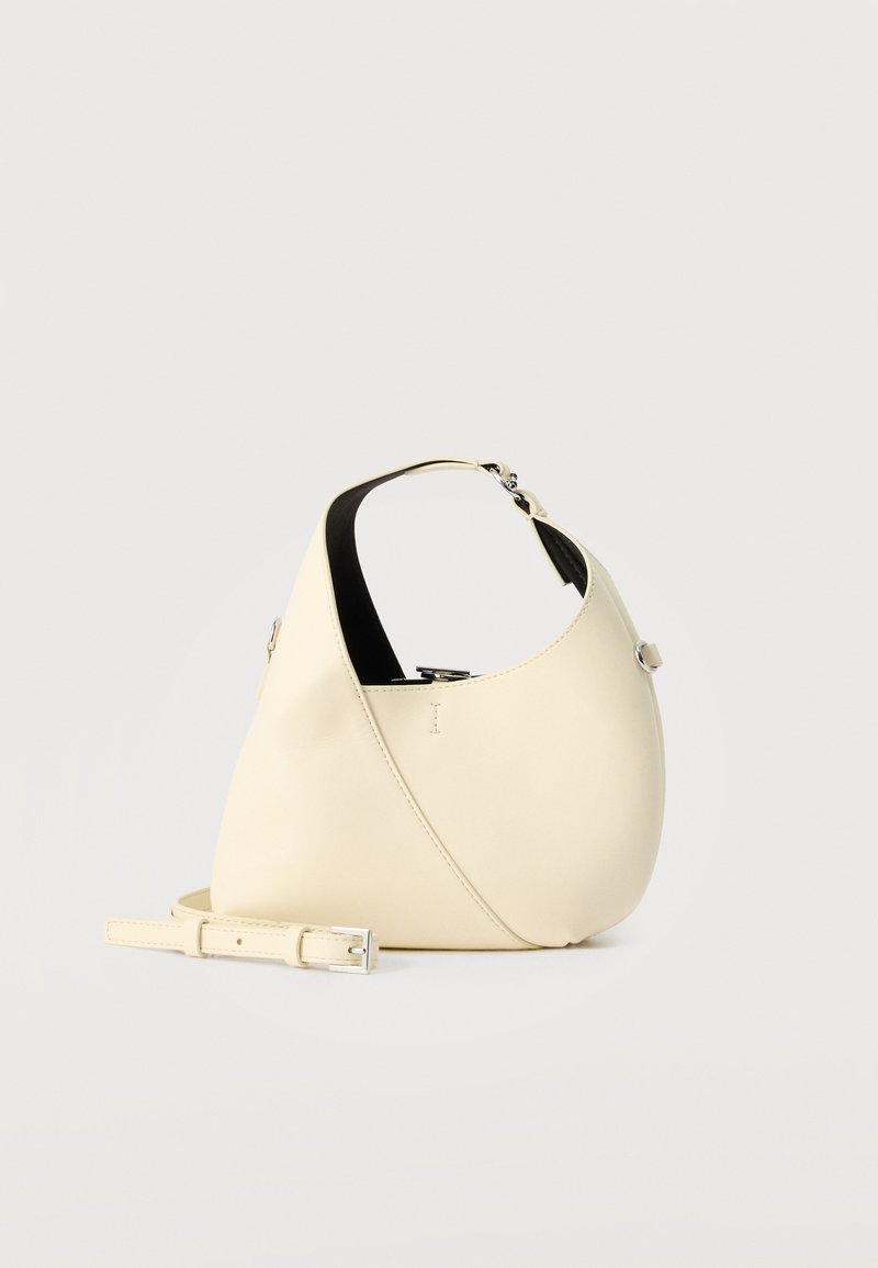 Κρεμ δερμάτινη τσάντα χόμπο με κοντή λαβή, ασημένια μεταλλικά στοιχεία και ρυθμιζόμενο αποσπώμενο ιμάντα ώμου σε λευκό φόντο.