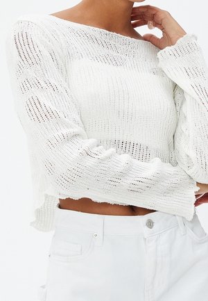Femme portant un crop top blanc en maille lâche à manches longues associé à un jean en denim blanc taille haute, main reposant près du menton.