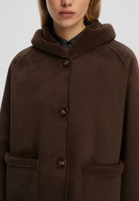 Cappotto marrone con cappuccio, dalla texture morbida, chiusura con bottoni e tasche applicate. Presenta un bordo in pile morbido e a contrasto sul cappuccio.