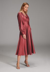 Bordeauxfarbenes Satin-Kleid mit tiefem V-Ausschnitt, langen Puffärmeln und geraffter Taille. Fließender A-Linien-Rock und silberne, schnürbare Absätze ergänzen den Look.
