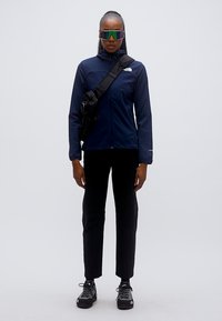 Μπλε σκούρο τζάκετ με φερμουάρ και ψηλό γιακά, μαύρο παντελόνι και αθλητικά παπούτσια. Το μοντέλο φοράει μεγάλα, ανακλαστικά γυαλιά ηλίου και τσάντα χιαστί.