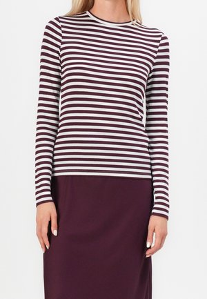 Vrouw draagt een maroon en wit horizontaal gestreept shirt met lange mouwen, gecombineerd met een effen maroon rok, tegen een effen achtergrond.