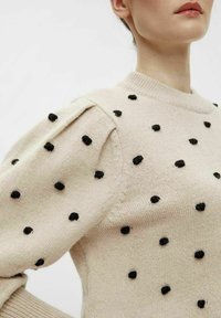 Striktrøje i lys beige med sorte hævede polka prikker; har pufærmer og ribhurtig halsudskæring. Blød tekstur.