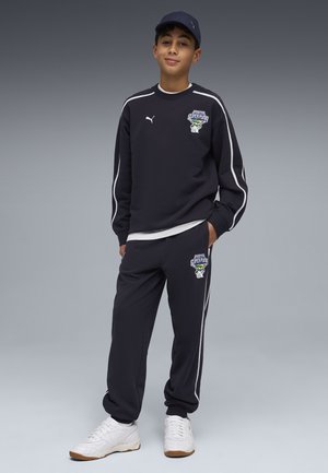 Jongen die een zwart Puma trainingspak draagt met witte strepen en logo's, witte sneakers en een zwarte pet, staand met één hand in de zak.