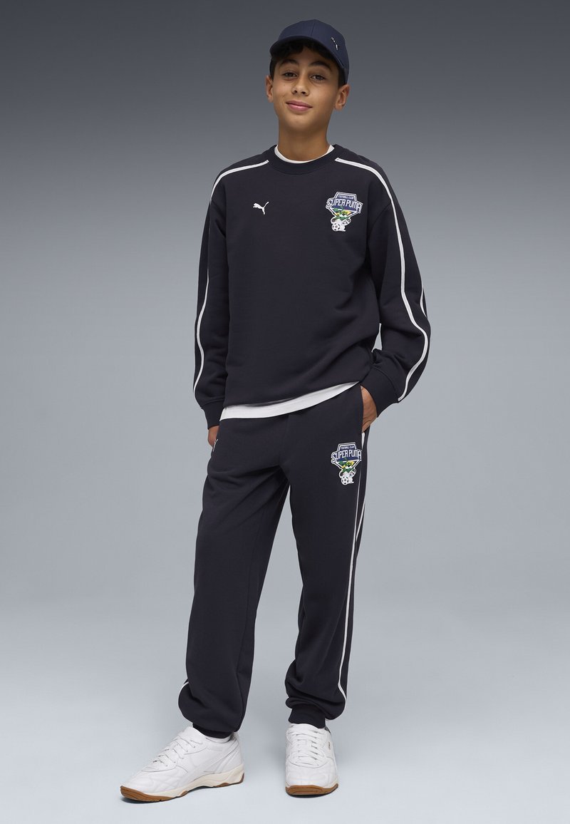 Garçon portant un survêtement Puma noir avec des rayures blanches et des logos, des baskets blanches et une casquette noire, debout avec une main dans la poche.