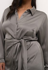 Chemise gris clair à boutons, en tissu lisse. Elle présente un col, des manches longues et un détail noué à la taille pour cintrer la silhouette. Surface texturée visible.